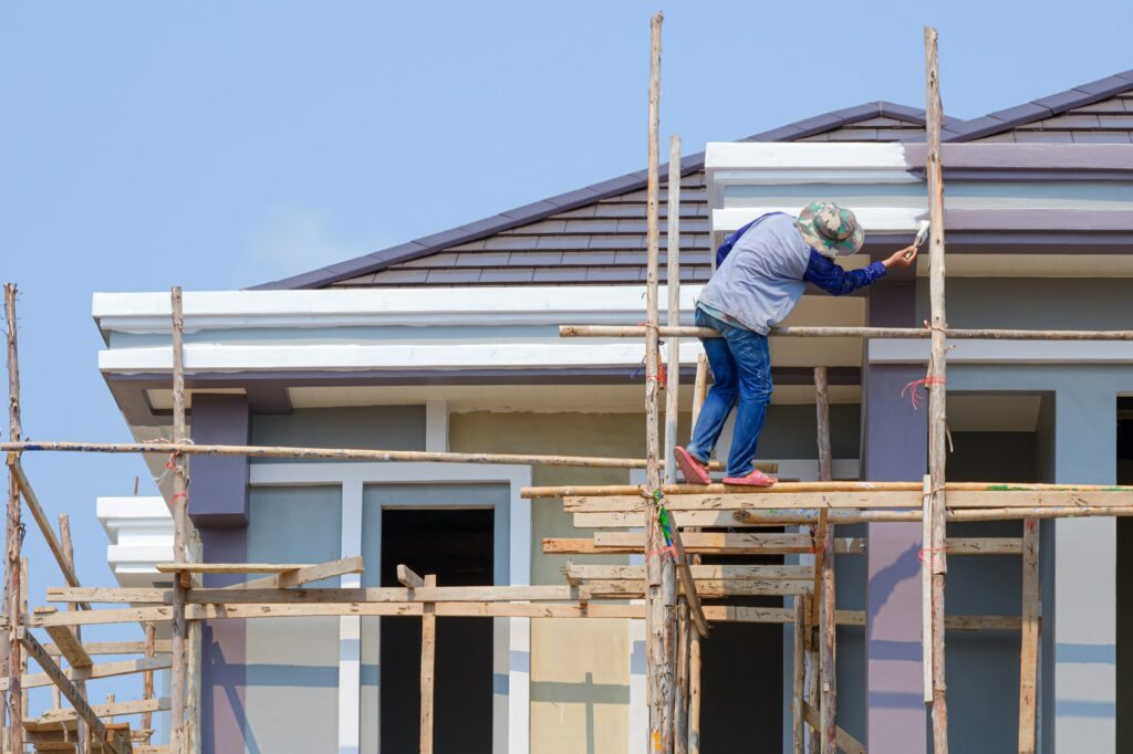 Painter on wooden scaffolding is painting roof eaves of modern house against blue sky background